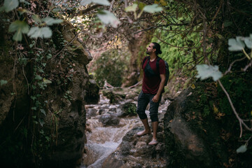 Naklejka na ściany i meble A young male tourist walks along a shallow tropical mountain river among the rocks.