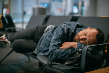 A tired male passenger sleeps on the seats waiting for boarding in the departure area of the...