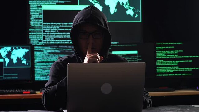 Asian Male Hacker In A Hood Showing Shh Gesture With Index Finger Near Mouth While Works On A Laptop With Maps And Data On Display Screens Of Multiple Monitors In A Dark Office Room

