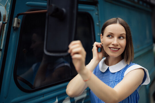 Pretty smiling woman in a retro blue dress with white collar looking in the mirror of a vintage bus