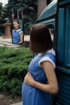 Pretty pregnant smiling woman in a retro blue dress with white collar looking in the mirror of a vintage bus