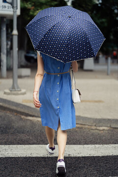 Back view of a woman, in a blue dress, with a white purse and an umbrella passing the crosswalk