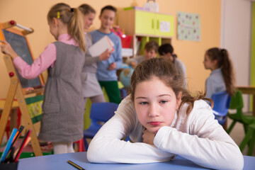 Upset girl sitting at table in schoolroom during break on background with other pupils