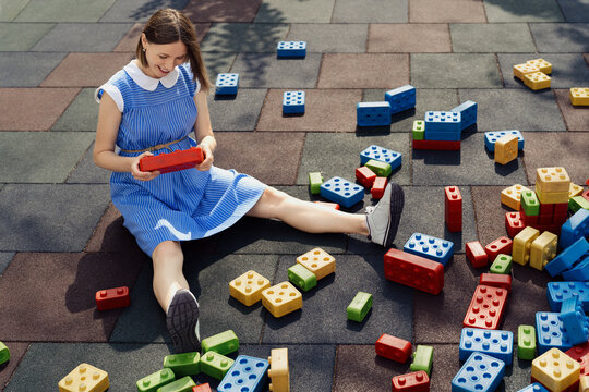 A beautiful pregnant woman in a blue dress playing with big colorful cubes on a children playground while sitting and watching the cubes. Pregnancy creative concept