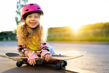 little beautiful girl with long blonde hair in a helmet with skate outside in summer, child...