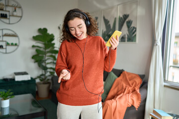 Happy pretty young woman wearing headphones using smartphone dancing at home. Cheerful girl listening music on mobile phone, singing song feeling relaxed standing in modern living room interior.