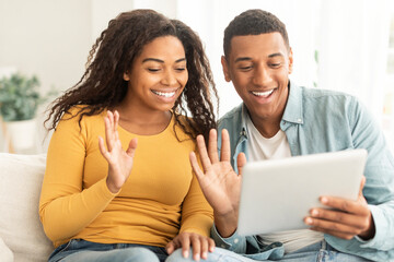 Cheerful young african american guy and lady waving hands at tablet webcam, greeting, say hello on sofa