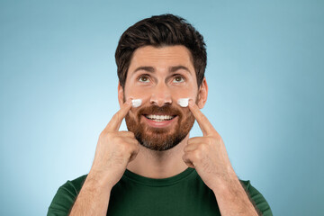 Bearded handsome man applying face cream on cheeks, standing on blue studio background, looking and smiling at camera