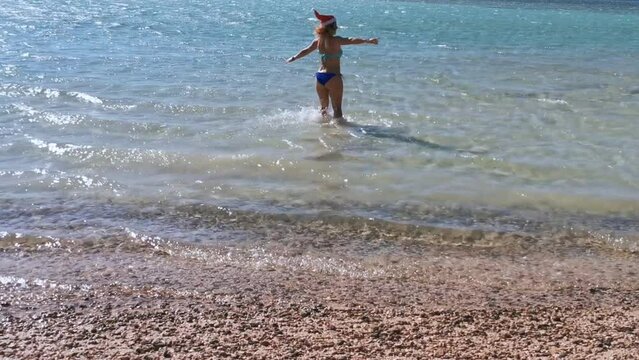 a girl in a blue swimsuit in a red santa hat walks on azure water on a summer sunny day. the girl radiates happiness and enjoys life and leisure. christmas celebration