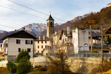View of residential houses and bell tower of parish church of small Alpine township of Finero located in Cannobina valley on background of snowy mountain ridge on winter day, Piedmont, Italy..