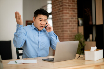 Fototapeta na wymiar Displeased mature asian man talking on cellphone while working on laptop, having problem with business communication