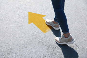 Planning future. Woman walking to drawn mark on road, closeup. Yellow arrow showing direction of way - obrazy, fototapety, plakaty