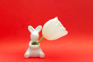 White ceramic figurine of Easter bunny on red background holds white tulip in its hands. Happy Easter greeting card. copyspace. Space for text. Horizontal banner. Religious holiday celebration concept