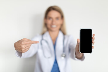 Medical Ad. Smiling Female Doctor Wearing Uniform Pointing At Blank Smartphone