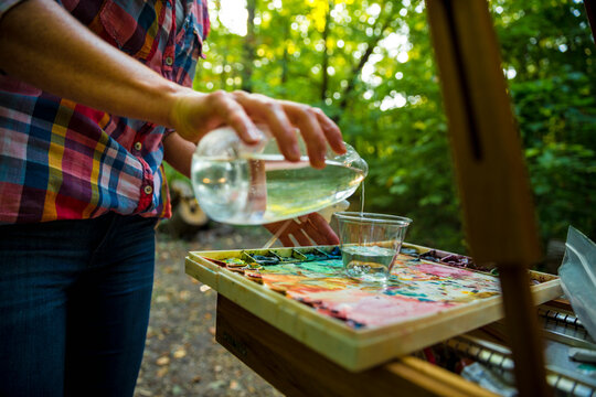 Female painter pouring water to cup standing on palette before painting in forest, Neenah, Wisconsin, USA