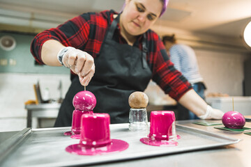Female baker wearing black apron glazing spherical mousse desserts with pink glaze. Professional dessert-making process. Horizontal indoor shot. High quality photo