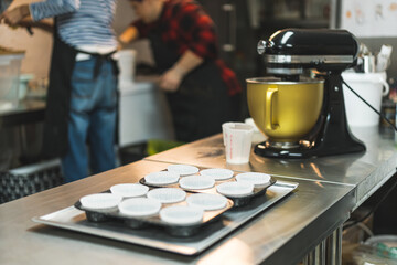 Cupcake tray with paper liners next to stand mixer machine with two bakers in black apron working in the background. Professional baking process. Horizontal indoor shot . High quality photo