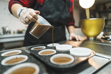 Female professional baker wearing black apron pouring out chocolate cupcake batter into paper cupcake liners in cupcake tray. Bakery. Horizontal indoor shot. High quality photo
