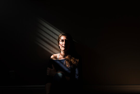 Portrait of woman sitting in a patch of striped light in a dark room.
