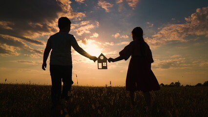 Children with their hands hold paper family house at sunset, sun shines through window. Symbol of home, happiness. Concept of building house for family. Dreaming of buying house. Boy girl play, park