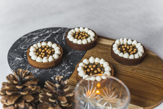 Close-up shot of four mini chocolate dessert tarts with golden sprinkles on wood and marble tray with lit candle and decorative pinecones. Festive baking. Horizontal shot. High quality photo