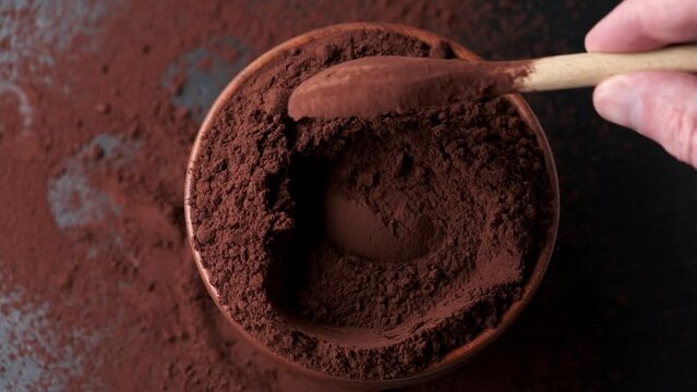 Spoon scooping cocoa powder from bowl on dark background.