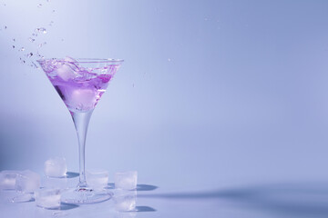 Cocktail splashing out of martini glass near ice cubes on light blue background, space for text