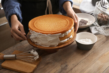 Woman smearing sides of sponge cake with cream at wooden table, closeup