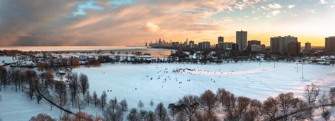 Wide angle aerial panorama of people sledding down Cricket Hill at sunset with bare trees and snow below and a bright yellow and pink sunset over the Chicago skyline beyond.