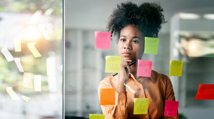 Young smiley attractive, businesswoman using sticky notes in glass wall to writing strategy business plan to development grow to success - obrazy, fototapety, plakaty