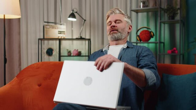 Bearded Man Sitting On Couch Closing Laptop Pc After Finishing Work In Living Room At Home Couch. Mature Guy Freelancer Working Online, Remote Job. E-learning, Browsing Internet On Notebook Computer