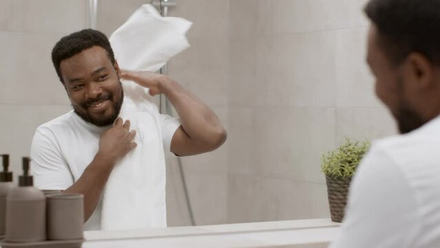 Young positive african american guy looking at mirror and smiling to himself at bathroom after washing in morning