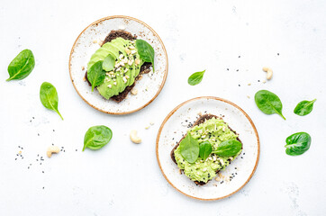 Avocado sandwich or toast on rye bread with spinach, crushed cashew nuts and sesame seeds, on plate, white table background, top view