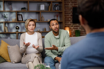 Fototapeta premium Multiracial quarrel between husband and wife at a session with a psychotherapist. A visit to a family psychologist for a married couple. A doctor consults an unhappy married couple. Help psychologist.
