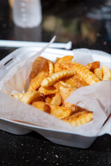 A view of a pile of crinkle cut french fries and potato wedges, in a styrofoam to go container.