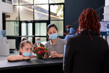Mother and child wearing protective face mask against covid19 while discussing medical insurance with receptionist during checkup visit examination in hospital lobby. Medicine service and concept