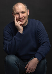 Lifestyle concept. Studio portrait of happy man with blue sweatshirt looking at camera with smile and holding one hand on cheek. Black studio background