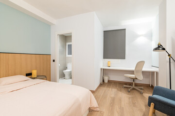View of bedroom with large bed, work area with white wooden table and comfortable leather office chair. Small private bathroom with open door. To side is comfortable armchair with floor lamp.