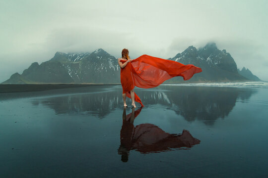 Woman in red dress with waving cape on beach scenic photography. Picture of person with hills on background. High quality wallpaper. Photo concept for ads, travel blog, magazine, article