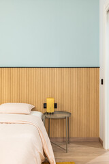 Vertical view of a bed with pillow with large warm duvet and linens in pleasant beige color. Modern design wall with wood paneling. On table is lamp with yellow frame for soft warm light at night.