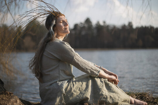 Close up woman in slavic pagan outfit sitting on lake coast portrait picture. Closeup side view photography with blurred background. High quality photo for ads, travel blog, magazine, article
