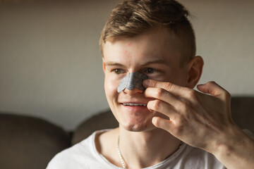 Smiling young man applying black nose patch to clean pore skin and remove acne or blackheads on nose. Skincare concept.