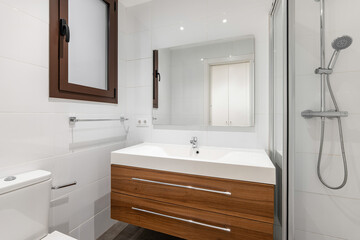 Bathroom with washbasin on hardwood vanity with two drawers for toiletries. Walls with white glossy tiles. Window is in plastic frame in dark wood color. Front door is reflected in mirror above sink.