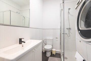 Bathroom with white walls and black marble floor tiles, toilet bowl and bucket for used toilet paper, vanity sink with shelves inside, shower area with glass partition and wash and dry washers.