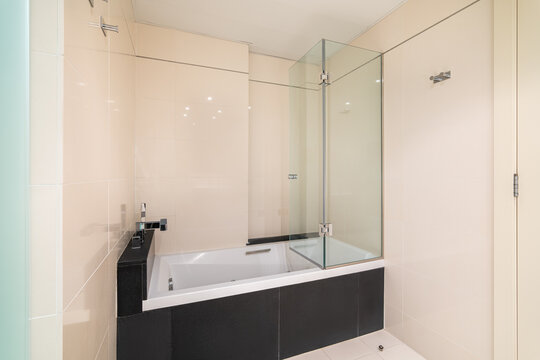 Bathroom With A White Bathtub For Bathing And Relaxation In Glossy Black Marble Tiles. Faucet Is Built Into A Ledge Above The Black Tile Bath. Glass Transparent Screen Protects From Splashes.