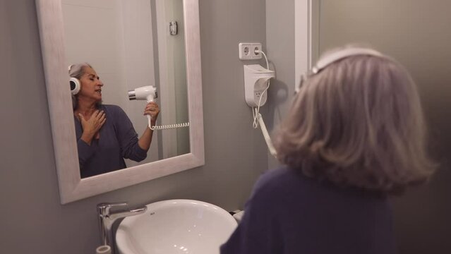 Proyecto sin títuloPortrait of senior woman wearing headphones and bathrobe indoors at home. singing in front of the mirror with the microphone dryer