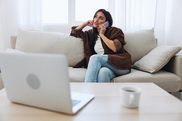 Obraz premium Woman blogger with phone lying at home and talking on the phone on sofa with laptop and working freelancer online, smiling with teeth