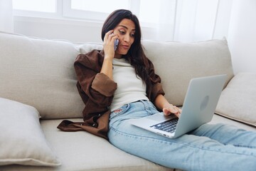 Woman blogger with phone lying at home and talking on the phone on sofa with laptop and working freelancer online, selfie