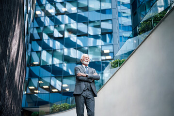 Low angle view of elderly professional looking away with arms crossed. Thoughtful senior businessman is standing outside modern business office building - Powered by Adobe