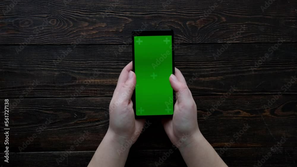 Wall mural Women's hands scroll the tape on the phone. Green screen. Top view of smartphone and wooden brown table